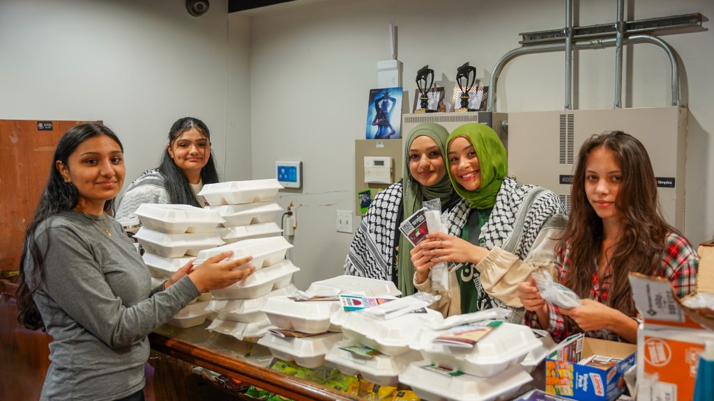 Five women grin while picking up styrofoam boxes of food from "Taste of Baladi." The third and fourth women are wearing hijabs. 