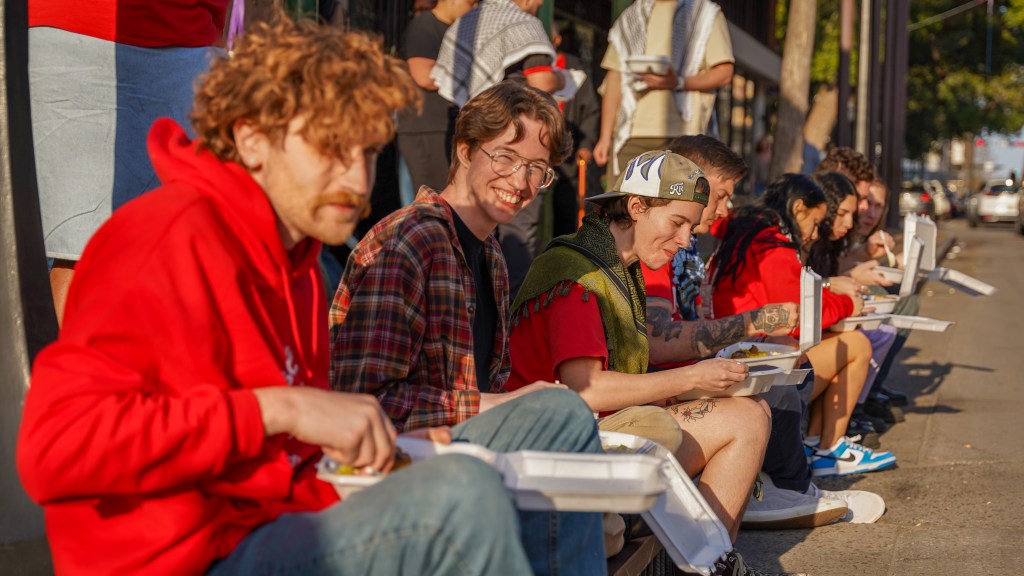 A group of people sit on a curbside to enjoy their food from "Taste of Baladi."