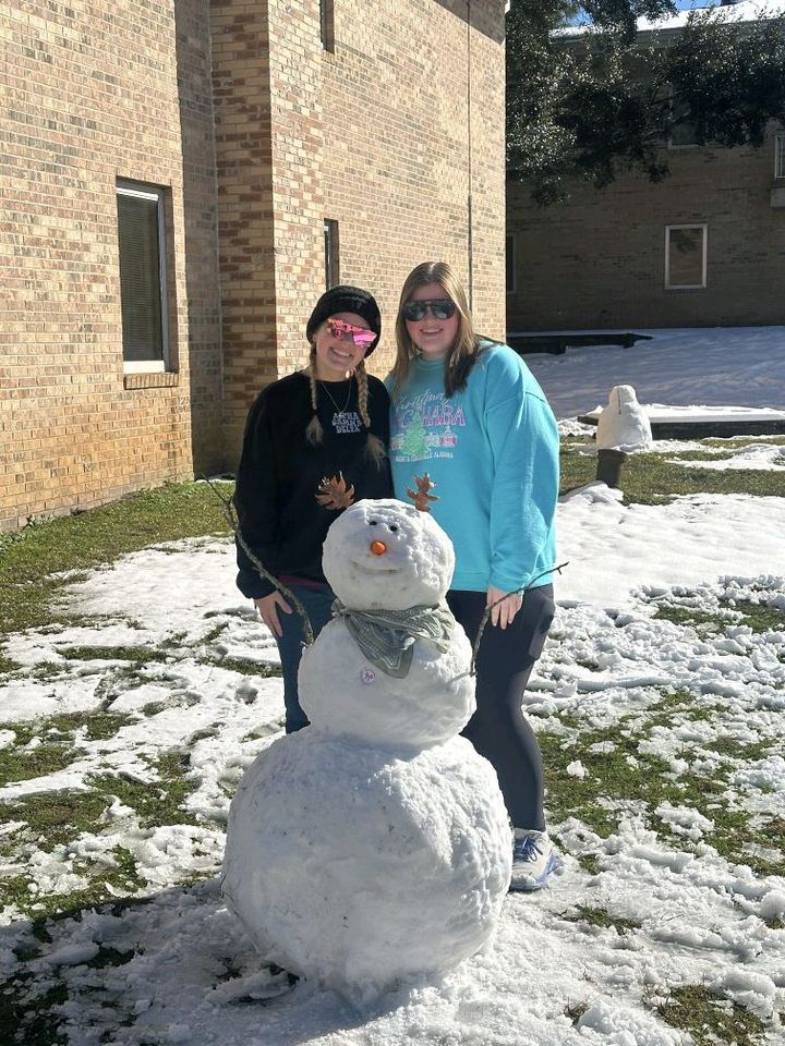 two average-height female students stand behind a snowman