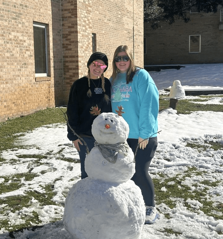 two average-height female students stand behind a snowman